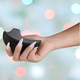 With long, manicured nails, a person holds an elegant Svakom Pulse Galaxie Air Pulse Stimulator in Midnight Black. The softly blurred pastel lights in the background enhance the serene atmosphere while the device promises discreet elegance with its gentle suction effect.